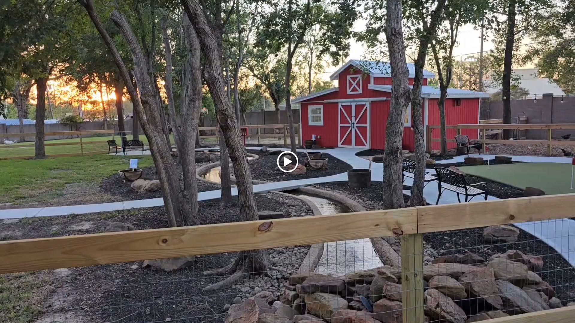 Outdoor garden area at sunset featuring a red barn-style building, winding paved walkways, benches, trees, a small water feature, and a putting green surrounded by a wooden fence.