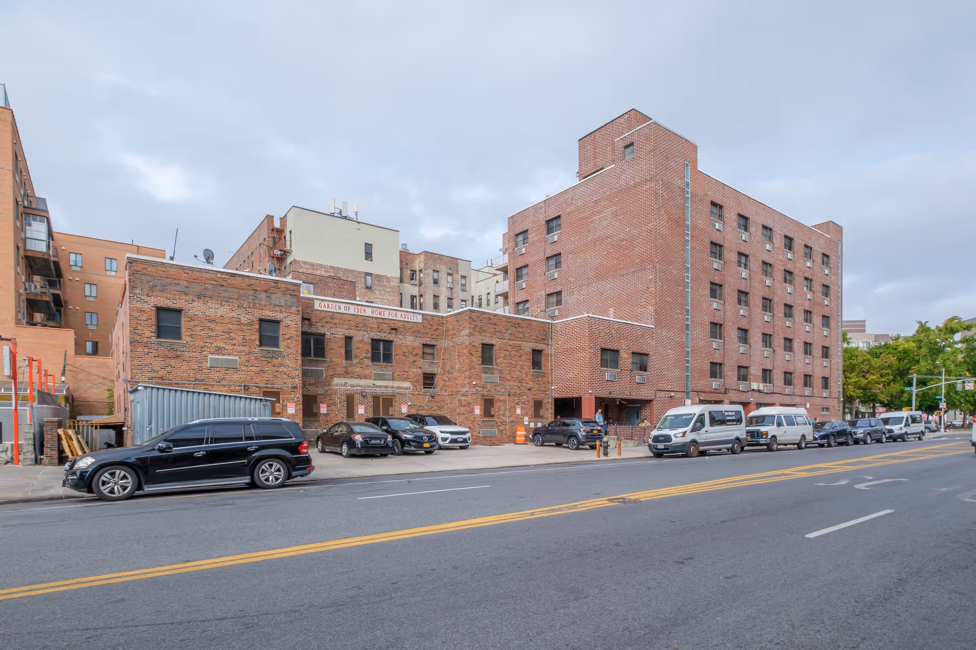 Exterior view of a multi-story brick building with a sign that reads 'Garden of Eden Home for Adults'. Several cars and vans are parked along the street and in a small parking area in front of the building. The sky is cloudy.