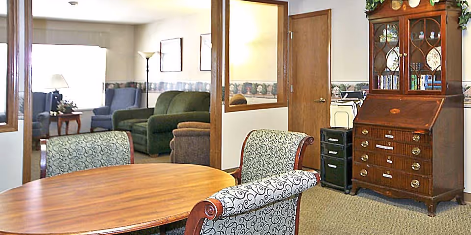 Sunlit senior living common room with a round wooden table and upholstered chairs in the foreground, sofas and armchairs in the background, and a wooden cabinet to the right.
