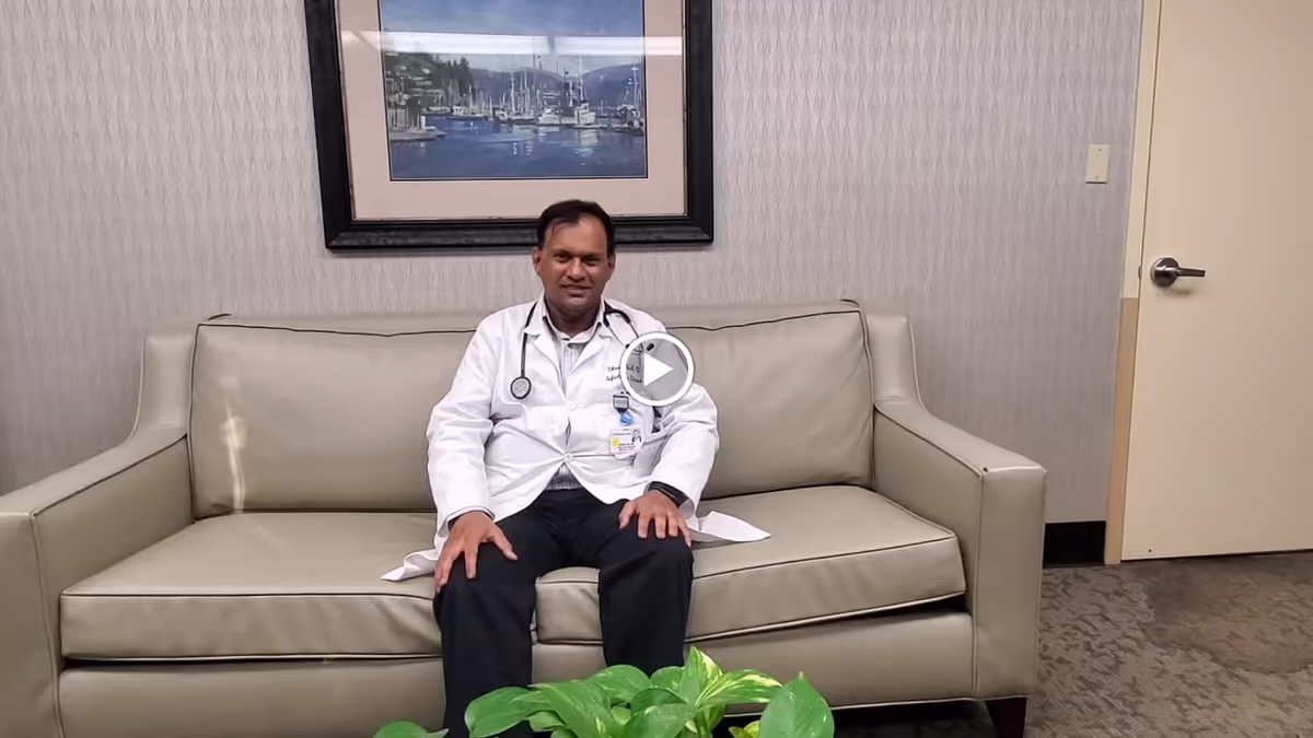 A man wearing a white medical coat and stethoscope sits on a beige leather couch in a room with patterned wallpaper. There is a framed picture of boats on the wall behind him and a green plant in the foreground. A closed door is visible to the right.