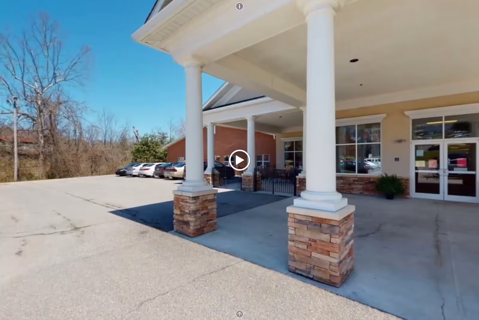 Covered front entrance of a nursing facility with large white columns on stone bases, a concrete drive and parked cars.