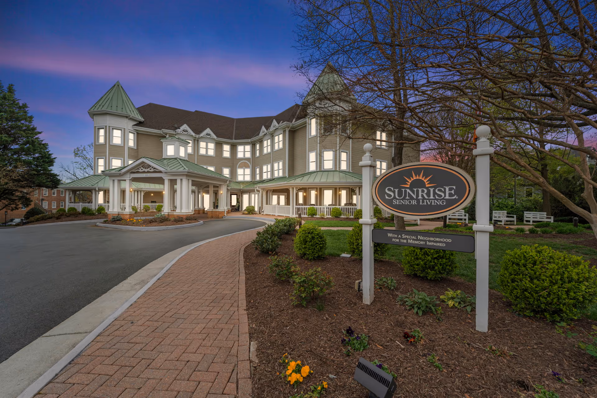 Exterior view of Sunrise Senior Living facility at dusk with a paved driveway, landscaped garden, and a sign that reads 'Sunrise Senior Living with a special neighborhood for the memory impaired'. The building has multiple windows and green roofs with a turret design.