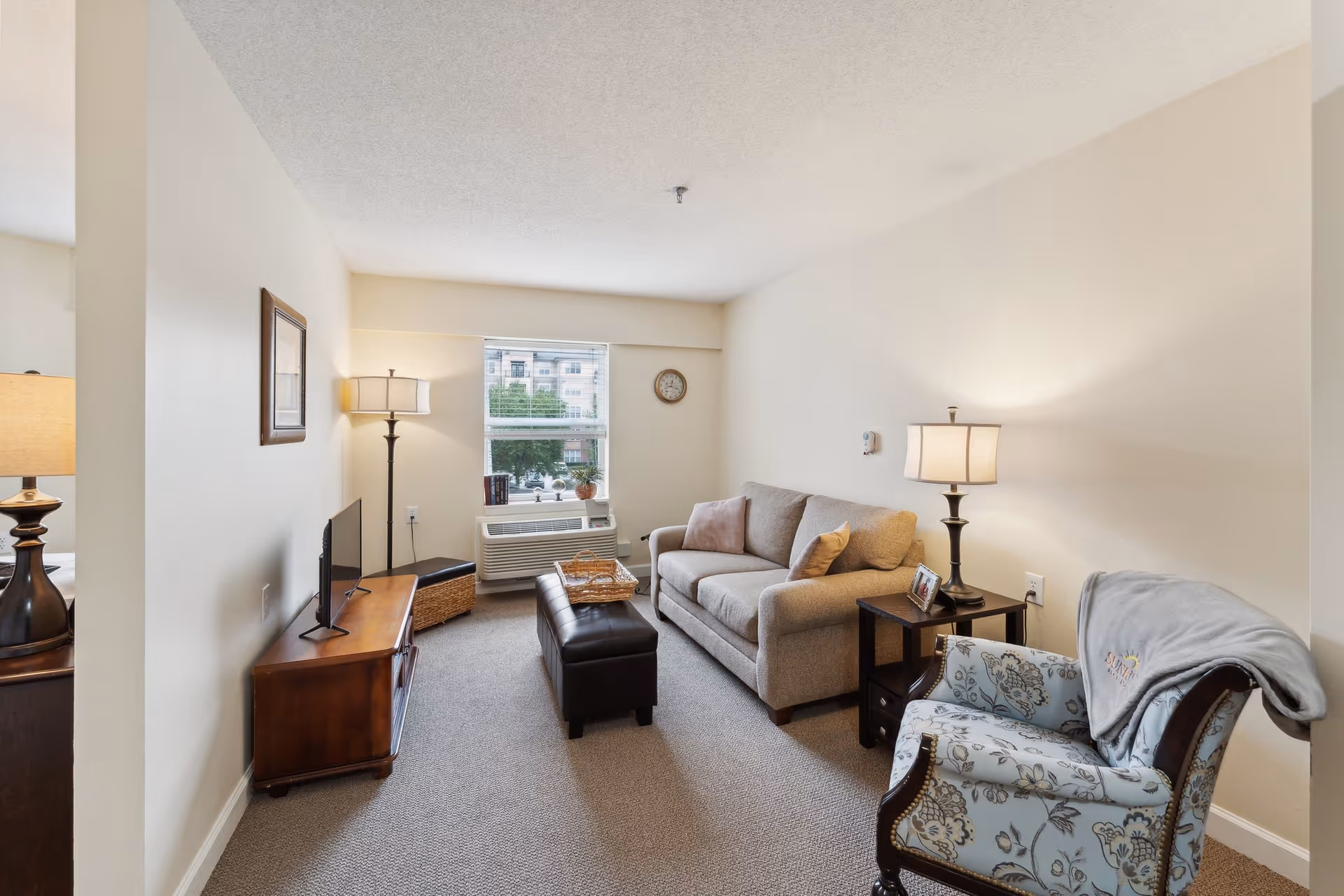Bright furnished living room with a sofa, patterned armchair, ottoman, TV stand and lamps beneath a window.