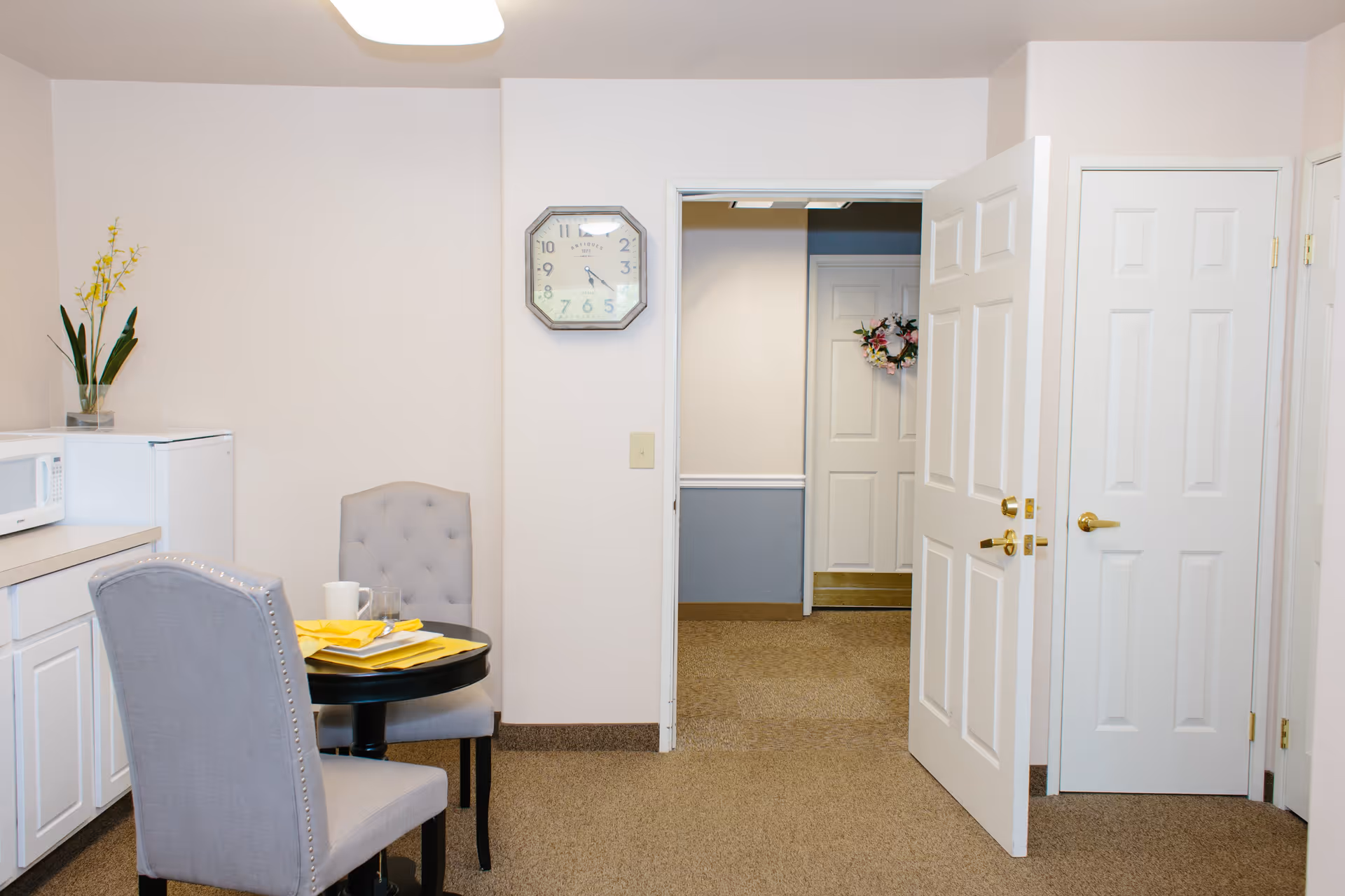 Small dining area with two gray upholstered chairs around a round black table set with yellow napkins, a white mug, and a glass. To the left, there is a white microwave on a countertop and a small white refrigerator below. A potted plant with yellow flowers is on the counter. The wall clock shows the time as 10:10. There are two white doors, one open and one closed, leading to a hallway with a door decorated with a floral wreath.