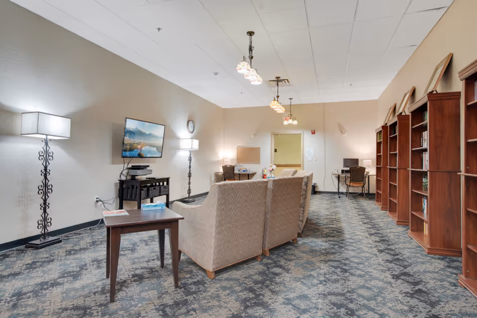 A spacious common area with patterned carpet flooring, beige walls, and ceiling lights. The room features a row of wooden bookshelves on the right, a seating area with beige armchairs and a small wooden table in the center, and a wall-mounted TV with a stand below it on the left. There are two floor lamps with decorative stands and a desk with a chair and computer in the back corner.