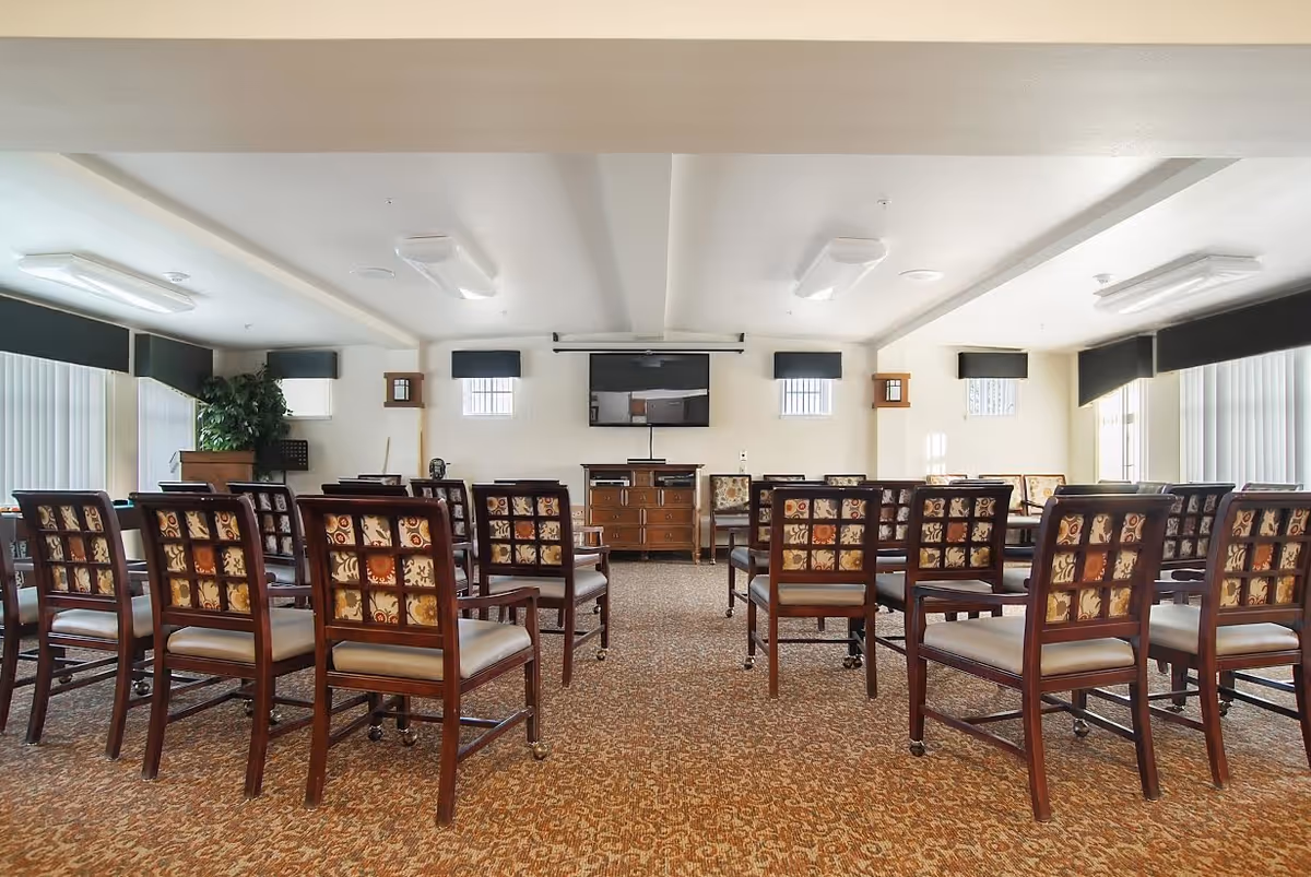 Light-filled activity/meeting room with rows of patterned wooden chairs facing a wall-mounted TV and media cabinet.