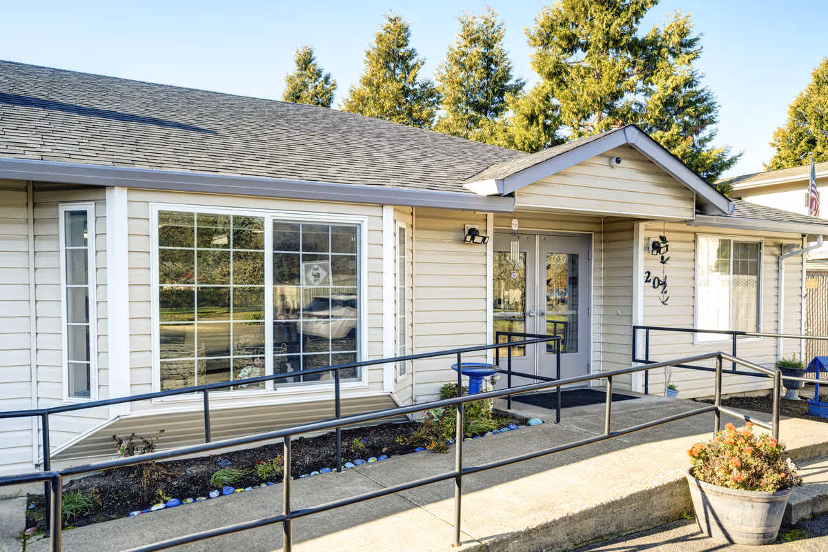 Exterior view of a single-story building with beige siding and a gray shingled roof. The building has large windows and a double glass door entrance with a small overhang. There is a concrete ramp with black metal railings leading up to the entrance, and a planter with flowers near the ramp. Trees and a clear blue sky are visible in the background.