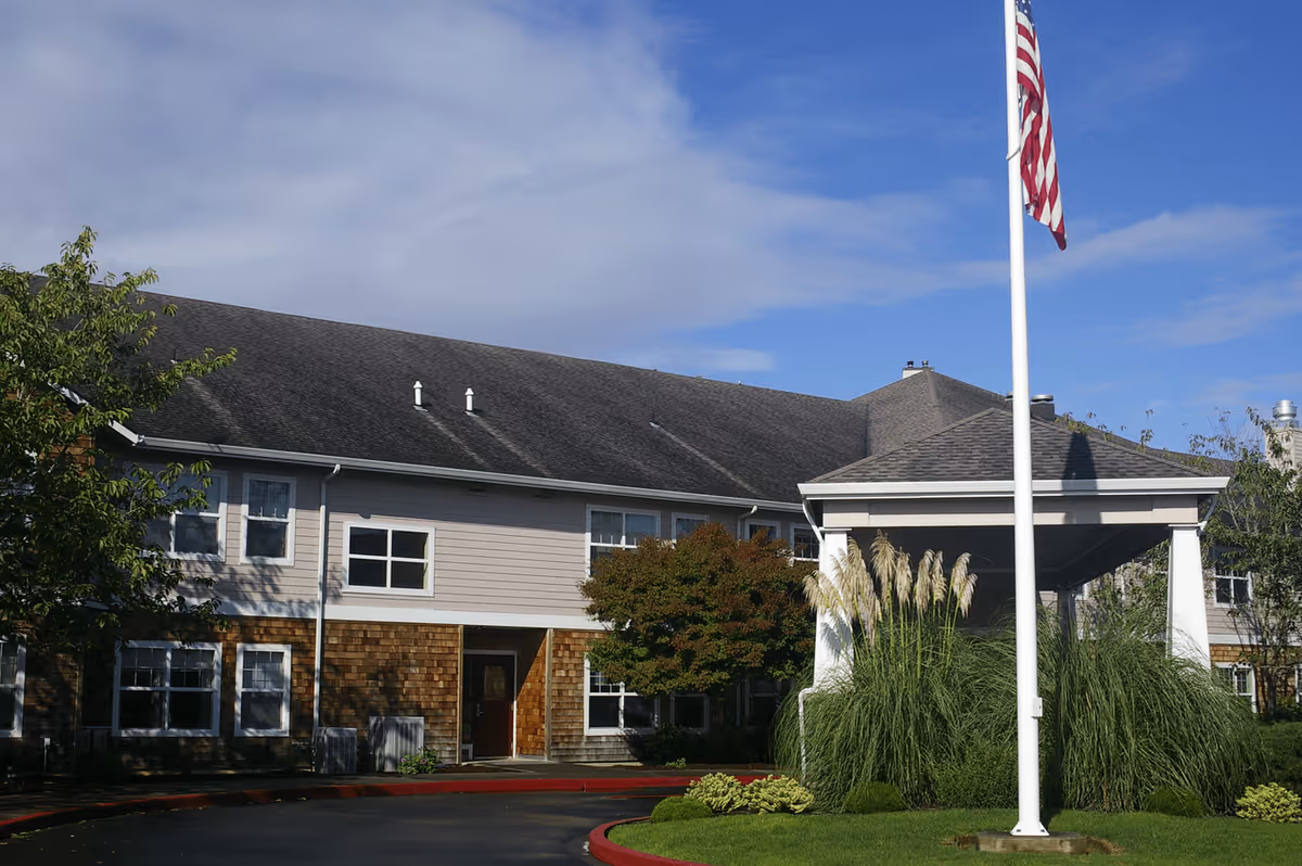 Exterior view of a two-story senior living facility building with a covered entrance, surrounded by greenery and a flagpole with an American flag in front under a partly cloudy sky.