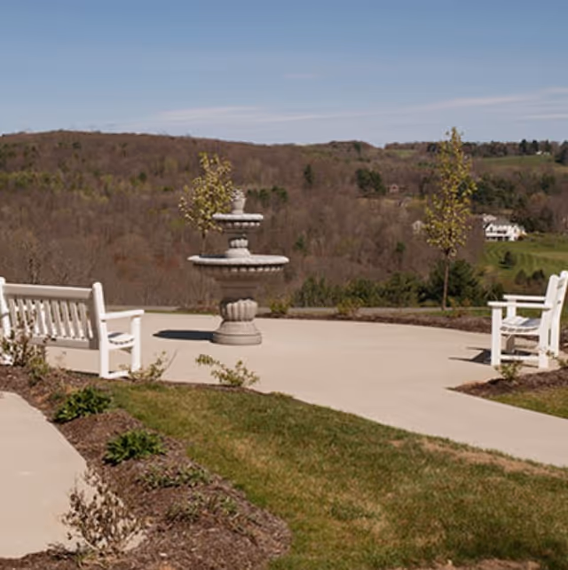 Outdoor patio area with two white benches facing a two-tier stone fountain, surrounded by small trees and landscaping, with a view of wooded hills and a distant house under a clear sky.