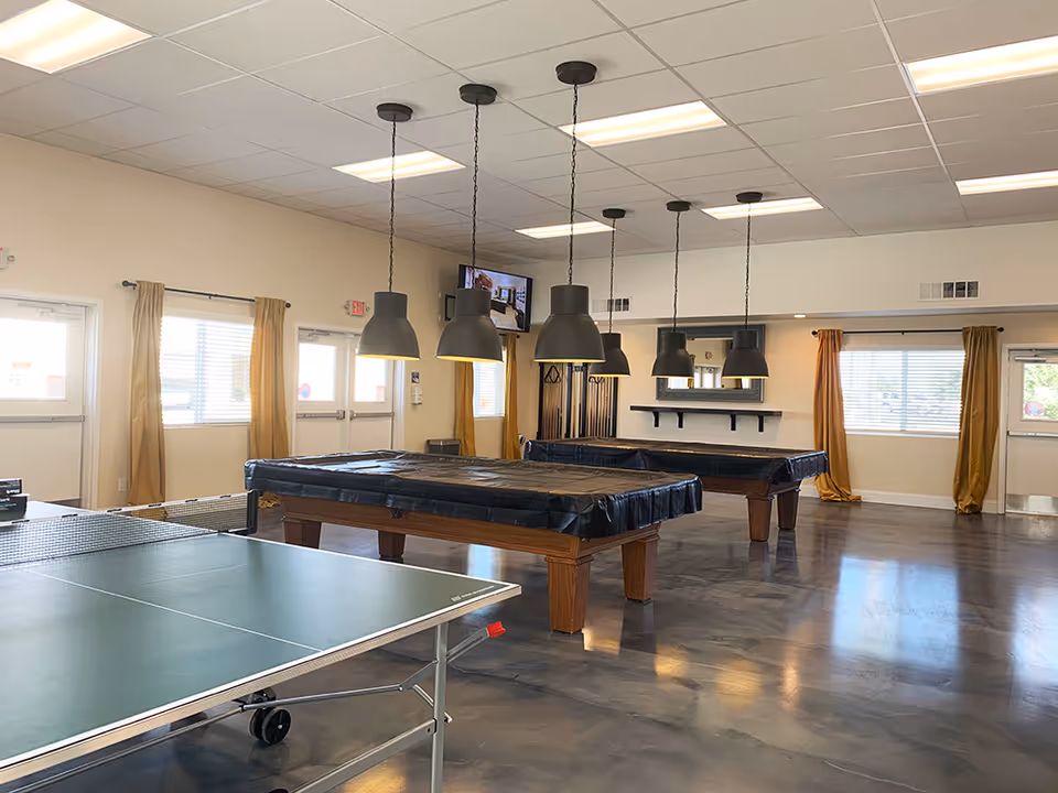 A spacious recreational room with two covered pool tables and a ping pong table in the foreground. The room has several large pendant lights hanging from the ceiling, windows with yellow curtains, and a wall-mounted TV. The floor is polished concrete, and there are multiple doors and windows letting in natural light.