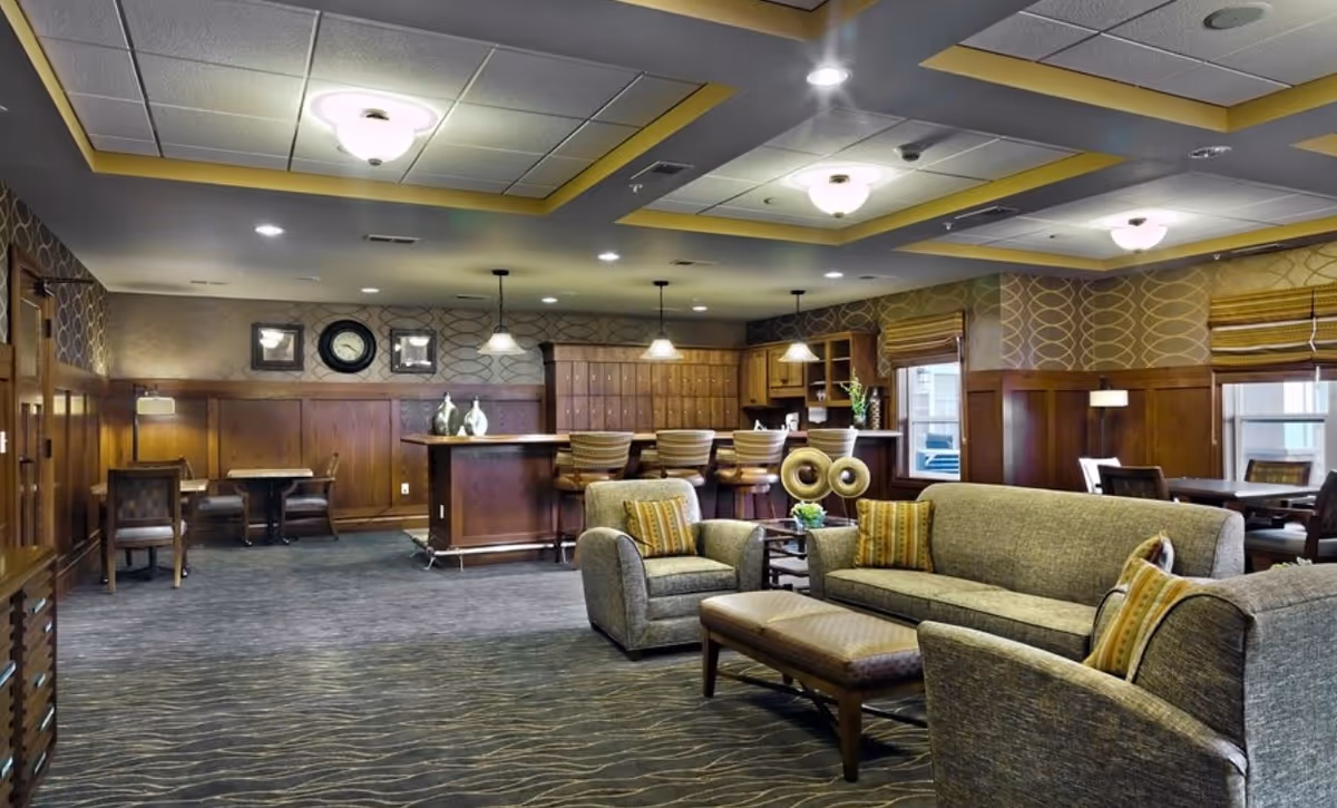 A spacious lounge area with comfortable seating including a sofa, armchairs, and a cushioned bench. The room features a carpeted floor, wood-paneled walls, and a ceiling with recessed lighting and decorative light fixtures. In the background, there is a bar counter with high chairs and pendant lights hanging above it. The room has windows with blinds and a few tables and chairs along the walls.