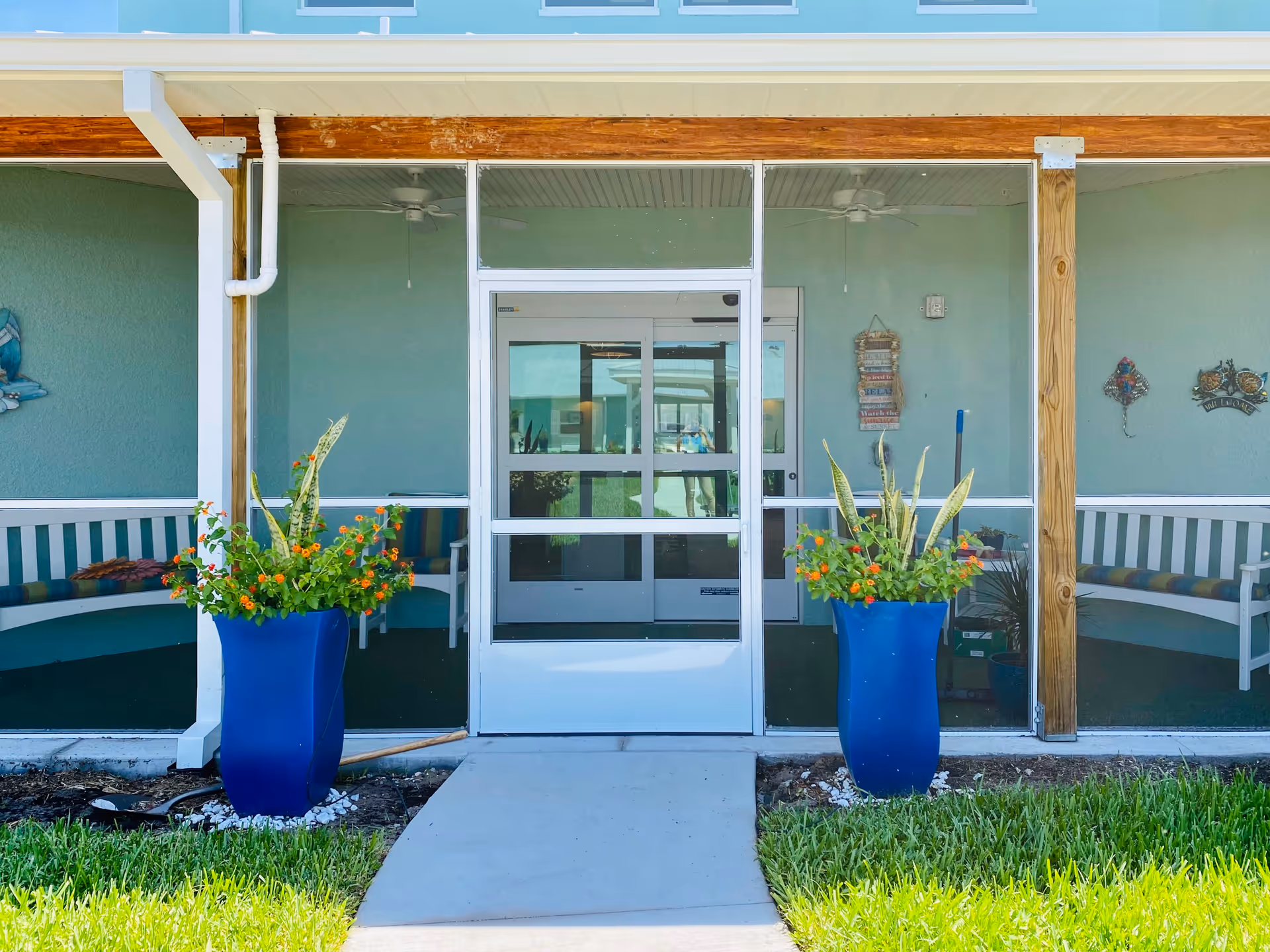 Entrance to a building with a glass door and screened porch area. Two large blue planters with green plants and orange flowers flank the walkway leading to the door. Inside the screened porch, there are white benches with colorful cushions and wall decorations.
