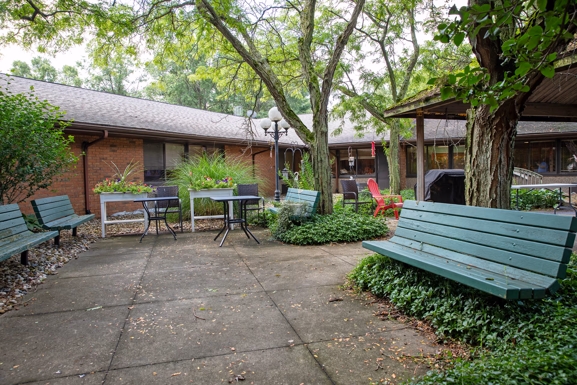 Outdoor courtyard area at East Lake Nursing and Rehabilitation Center with green benches, small tables and chairs, trees, plants, and a brick building in the background.