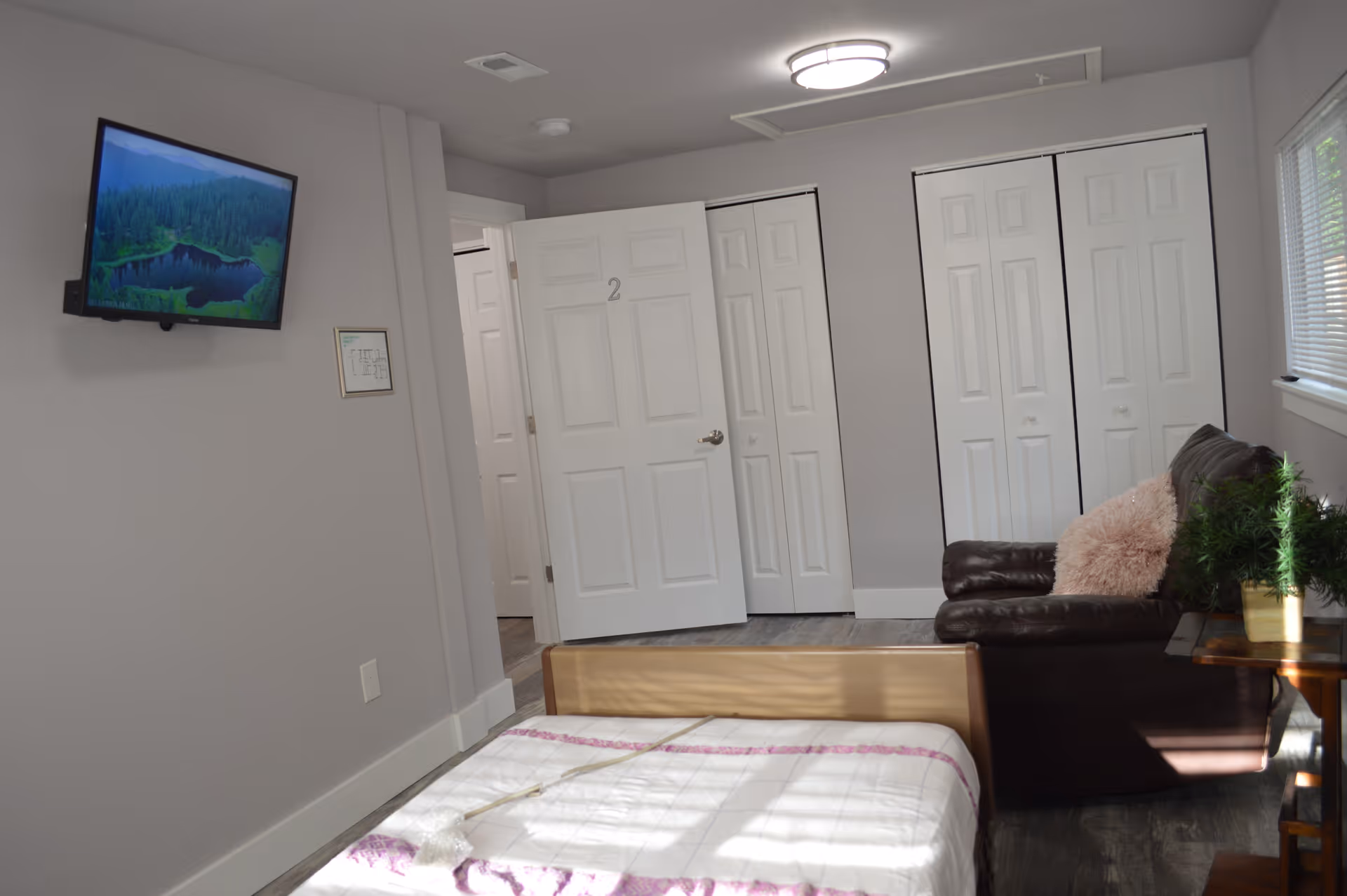Interior view of a senior living facility room with a bed in the foreground, a brown leather couch with a pink fluffy pillow, a small table with a plant, white closets, and a wall-mounted TV displaying a nature scene.