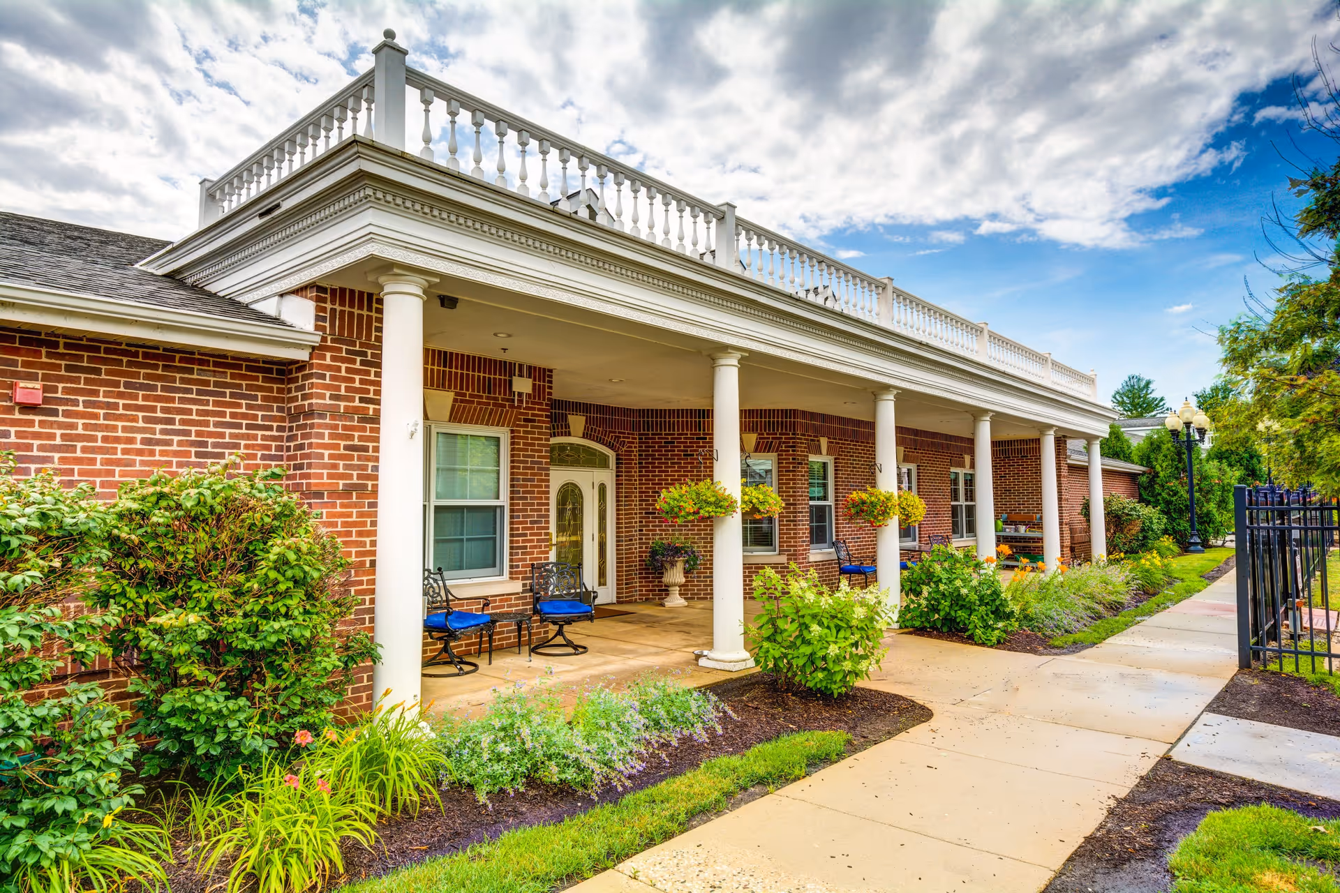 Exterior view of a brick building with white columns supporting a covered porch. The porch has several chairs with blue cushions and hanging flower baskets. There is a paved walkway leading alongside the building, surrounded by green shrubs and plants under a partly cloudy sky.
