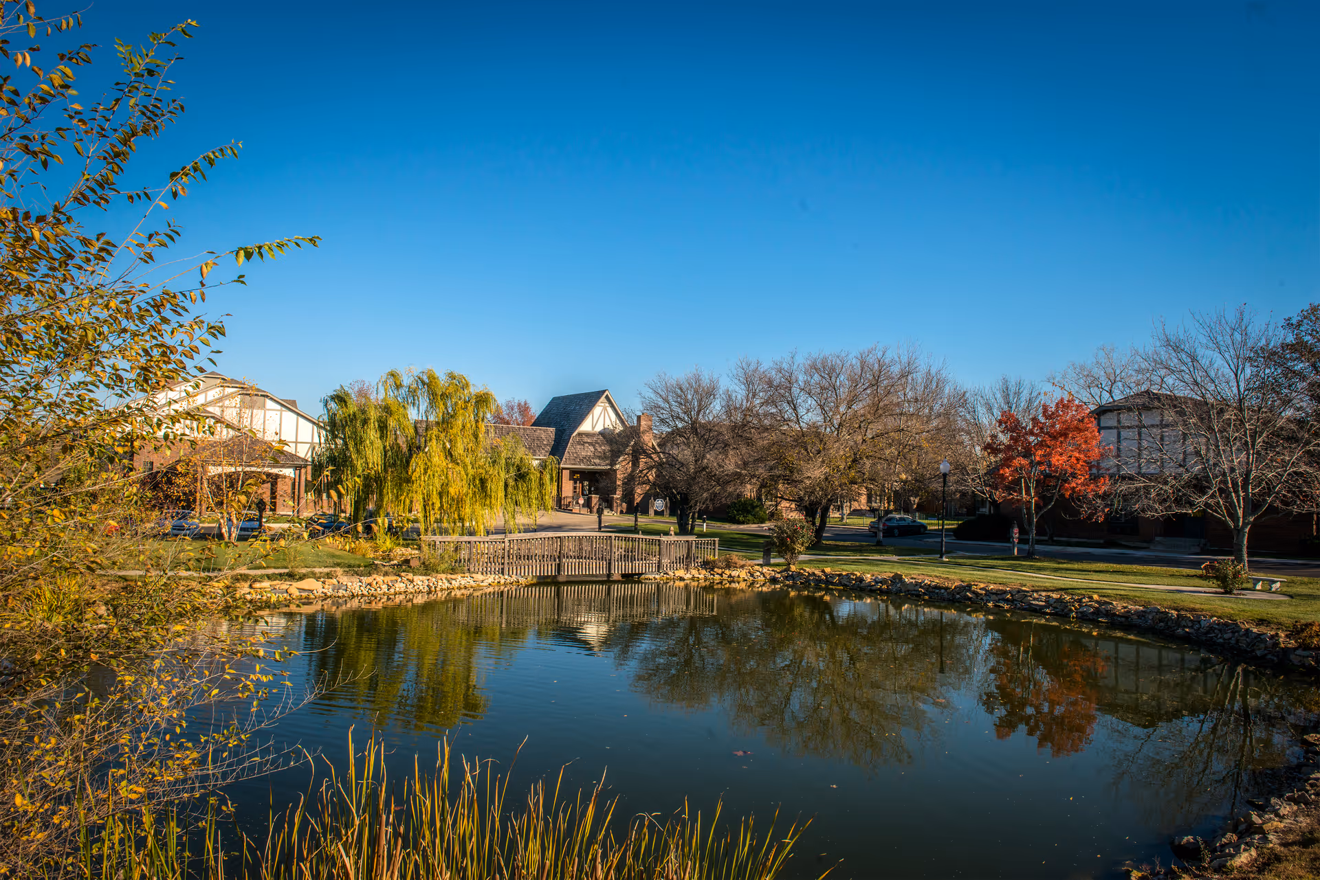 A landscaped pond with a small wooden bridge reflecting trees and nearby buildings under a clear blue sky.