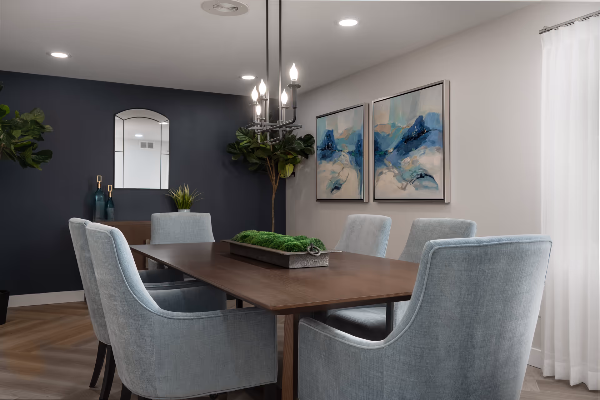 A modern dining room with a rectangular wooden table surrounded by six light blue upholstered chairs. The room features a dark accent wall with a decorative mirror and plants, two abstract blue and white paintings on a light-colored wall, a chandelier with exposed bulbs hanging above the table, and sheer white curtains allowing natural light to enter.