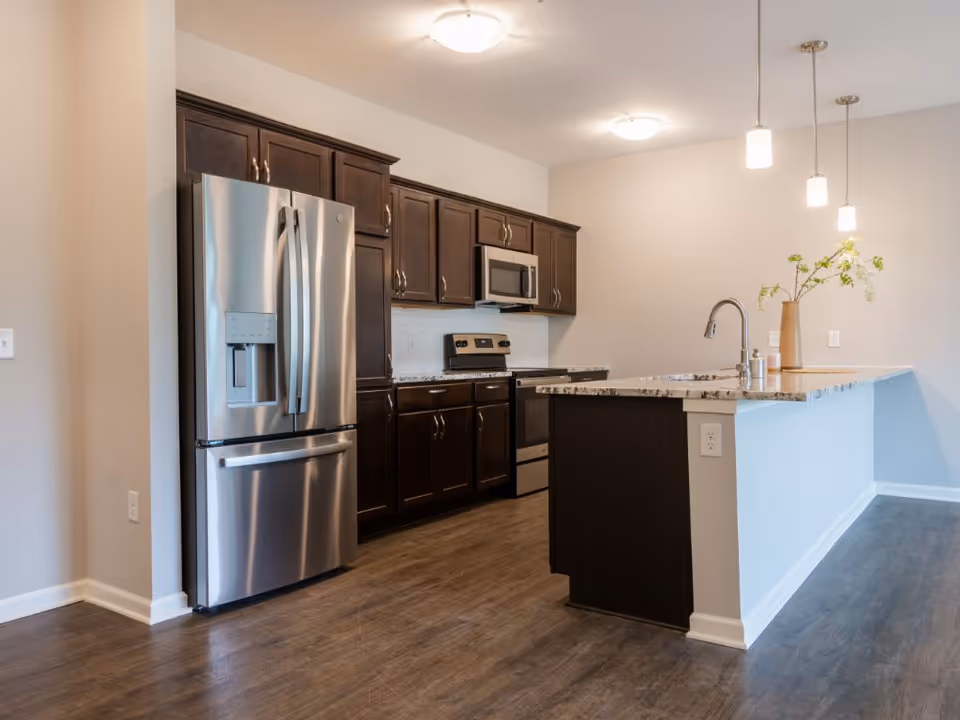 Modern kitchen with stainless steel refrigerator, dark wood cabinets, a granite-topped island, and pendant lights.