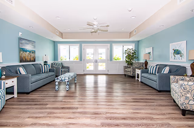 A spacious and bright living room area with light blue walls and wood flooring. The room features two gray sofas with striped and patterned cushions, two patterned armchairs, a small patterned bench, side tables with lamps, and framed artwork on the walls. Large glass double doors at the far end let in natural light.