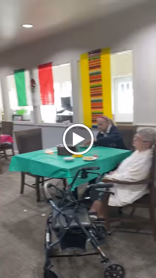 Two elderly women sitting at a table covered with a green tablecloth in a common area. One woman is using a walker positioned near her chair. The room has decorations including a Mexican flag and a colorful vertical banner on the wall behind them.