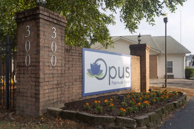 Entrance sign for Opus Post-Acute Rehabilitation facility mounted on a brick wall with the number 300 displayed on two brick pillars. There are flowers planted in front of the sign and a building with a gray roof in the background.