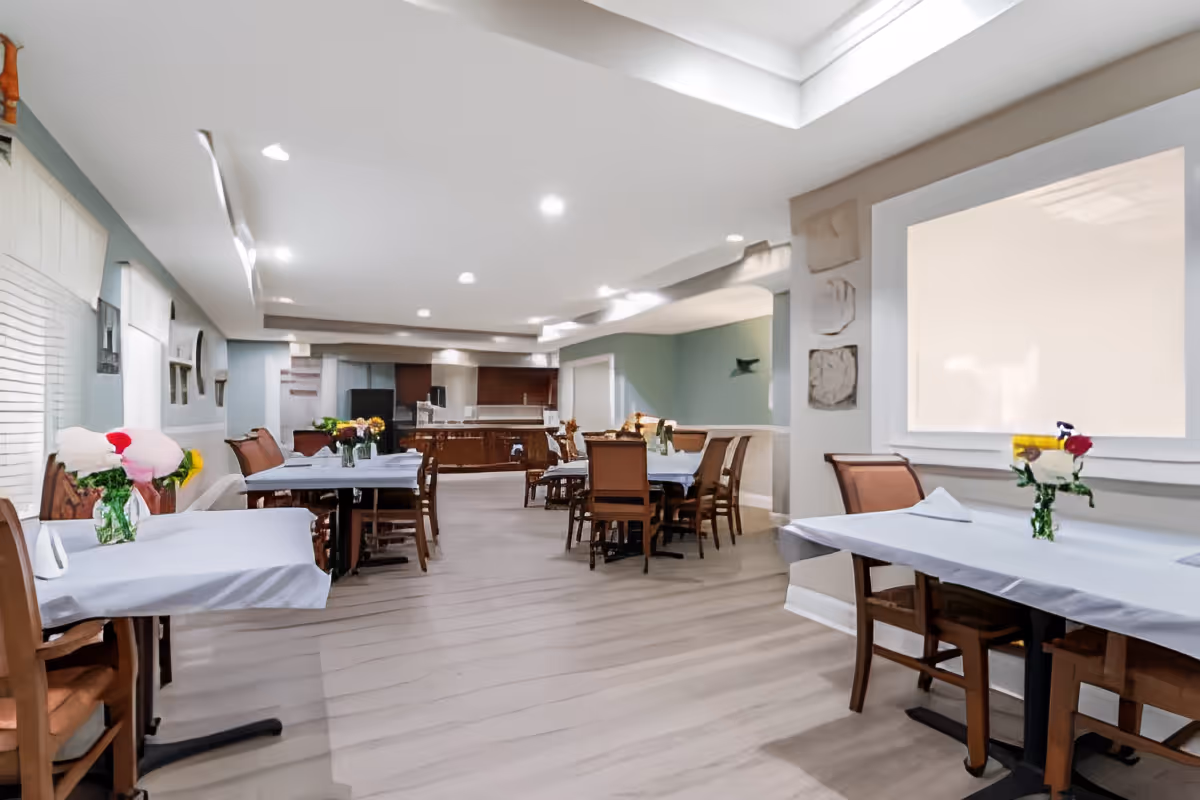 A bright and spacious dining room with several tables covered in white tablecloths, each adorned with small vases of colorful flowers. Wooden chairs are arranged around the tables. The room features light-colored walls, recessed ceiling lights, and a large window on the right side. The background shows a kitchen area with dark cabinetry.