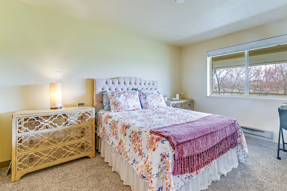 Bright bedroom with a floral quilted bed, tufted headboard, matching decorative nightstands with lamps, and a window showing trees outside.