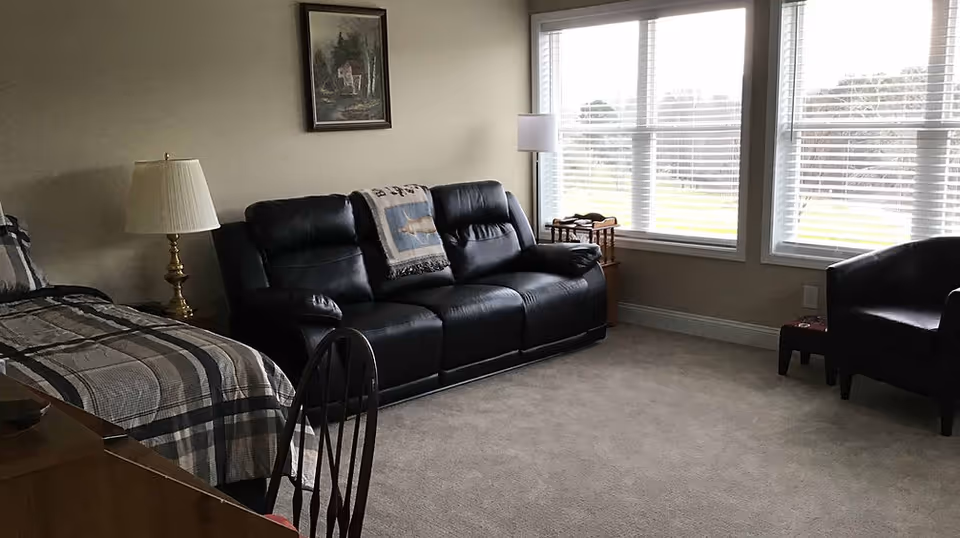 A senior living facility room with a bed on the left side covered in a plaid blanket, a black leather couch with a decorative throw blanket, a brass lamp on a side table, a wooden chair in the foreground, a small wooden table near large windows with white blinds, and a black armchair on the right side. The room has beige walls and carpeted floor.