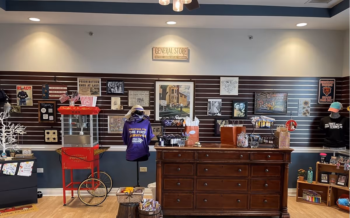 Interior view of a general store area in a senior living facility featuring a red popcorn machine, a wooden counter with various gift bags and items, mannequins dressed in themed shirts, and a wall decorated with framed pictures and signs.