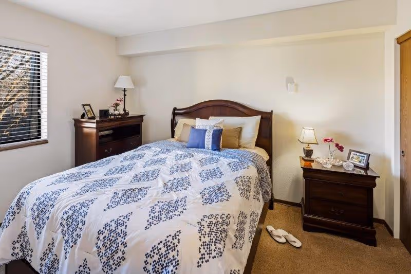 A cozy bedroom with a wooden bed frame and a white and blue patterned comforter. The bed has multiple pillows including a blue decorative pillow. There is a wooden nightstand on the right side with a small lamp, a framed photo, and a flower vase. On the left side, there is a wooden dresser with a lamp, a framed photo, and some decorative items. A window with blinds is on the left wall, and a pair of white slippers is on the carpeted floor near the nightstand.