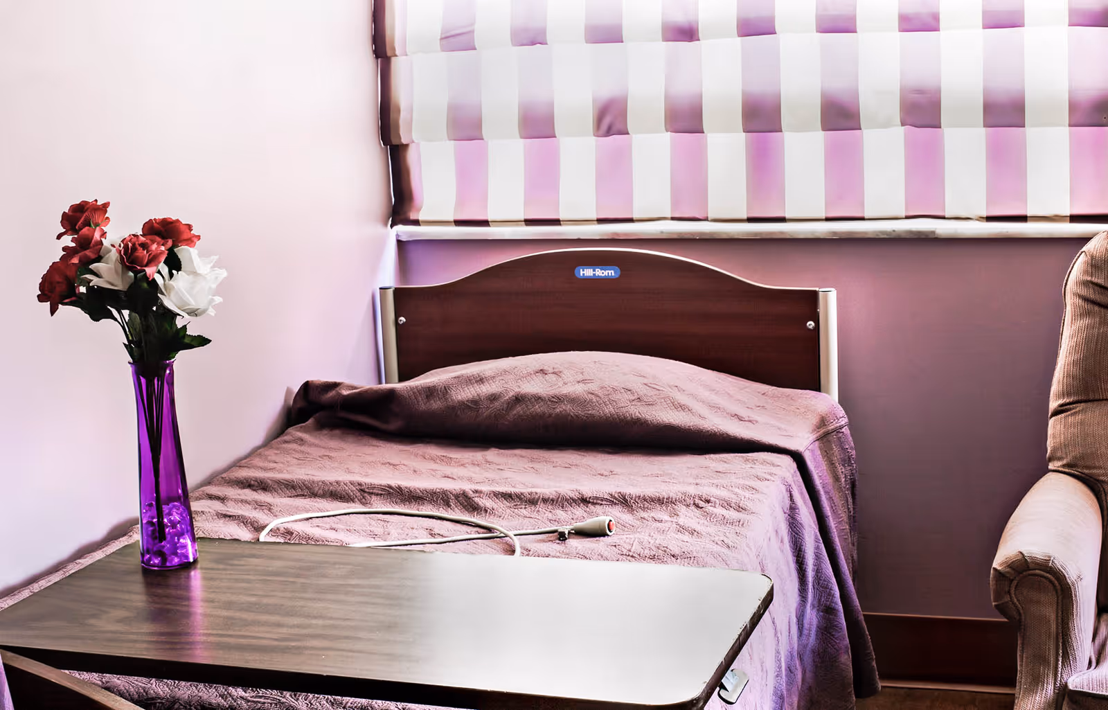 A small bedroom with a single bed covered in a purple blanket, a wooden headboard with a Hill-Rom label, a purple vase with red and white flowers on a table next to the bed, and a beige armchair partially visible on the right. The window has purple and white striped blinds.