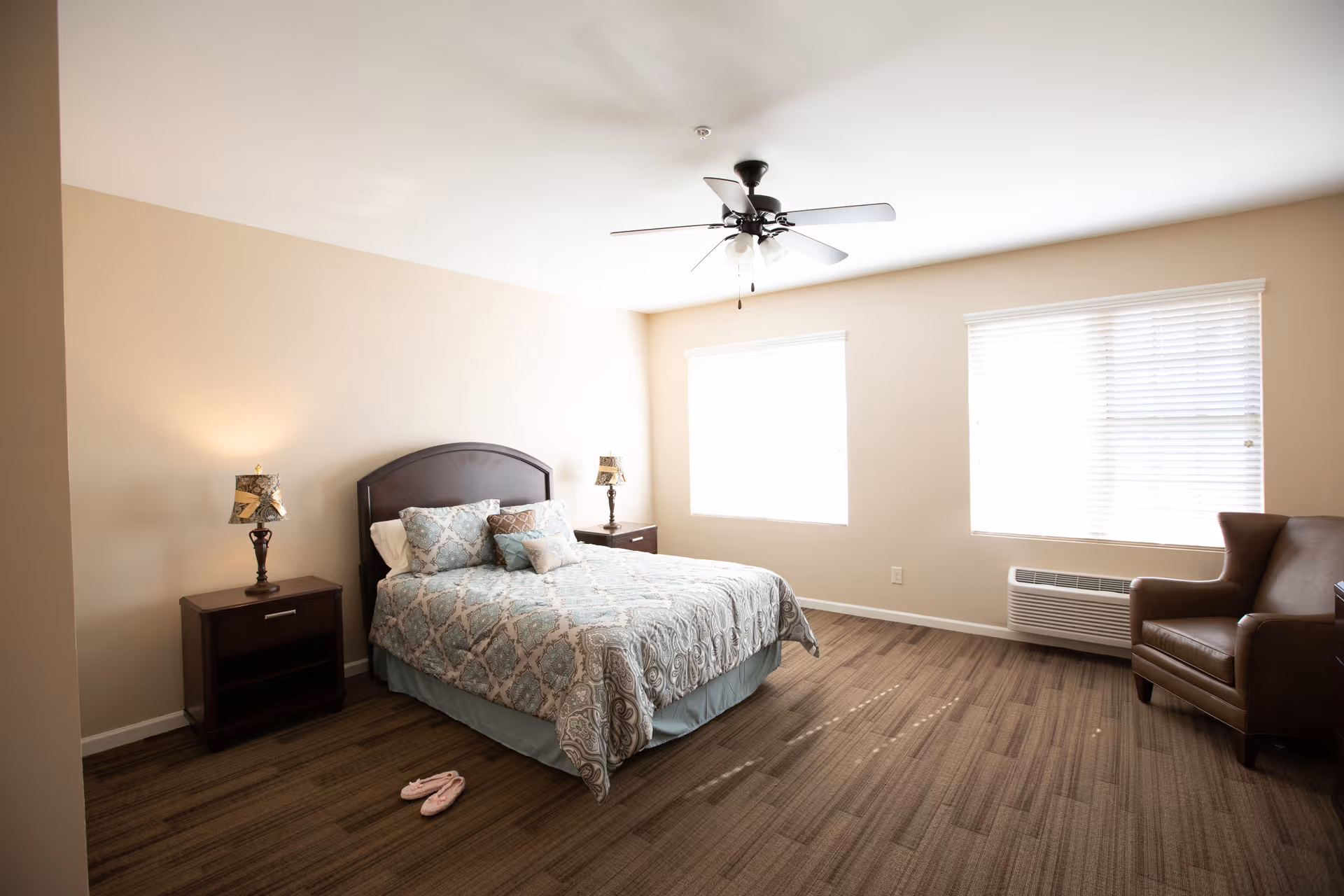 A well-lit bedroom with a double bed featuring patterned bedding and multiple pillows. There are two wooden nightstands with lamps on either side of the bed. The room has two large windows with blinds, a ceiling fan with lights, a brown armchair, and carpeted flooring. A pair of pink slippers is placed on the floor near the bed.