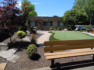 Courtyard with a wooden bench, small putting green, landscaped walkway and shrubs in front of a low single-story building.