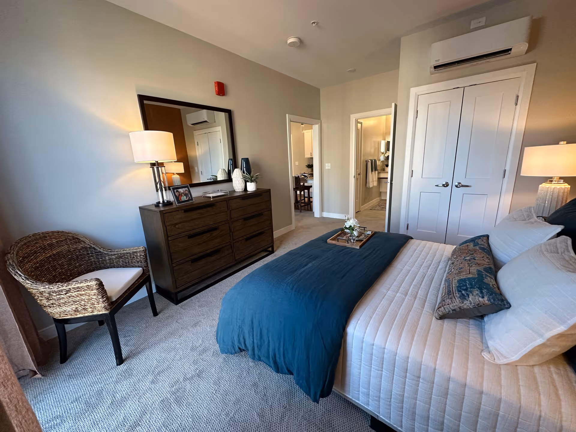 A neatly arranged bedroom with a bed covered in white and teal bedding, a wooden dresser with a large mirror above it, a wicker chair with a cushion, and two doors leading to other rooms. The room has beige walls and carpeted flooring, with a lamp on the dresser and another on a bedside table.