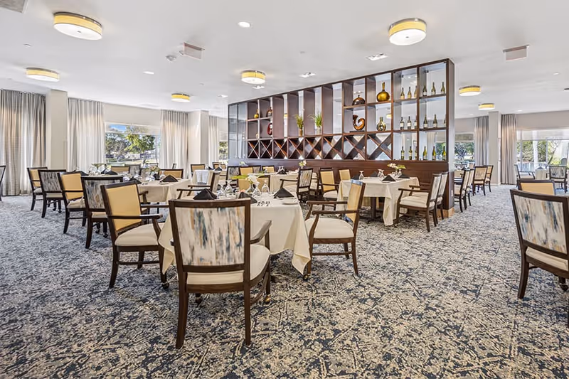 A spacious dining room with multiple tables covered in white tablecloths, each set with glassware and napkins. The room features patterned carpet flooring, wooden chairs with cushioned seats and backs, large windows with curtains allowing natural light, and a decorative wooden shelving unit displaying bottles and vases.