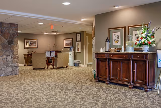 Carpeted senior living common area with seating, a wooden buffet holding coffee and a flower arrangement, and framed artwork on the walls.