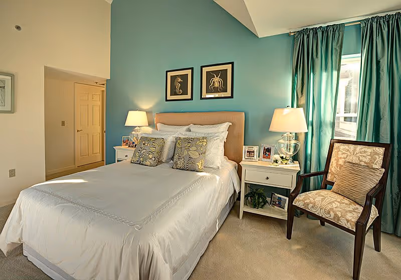 A neatly made bed with white linens and decorative pillows in a bedroom with teal accent wall. Two framed sea creature artworks hang above the bed. There are two white nightstands on either side of the bed, each with a lamp and framed photos. A patterned armchair with a cushion is placed near a window with teal curtains.