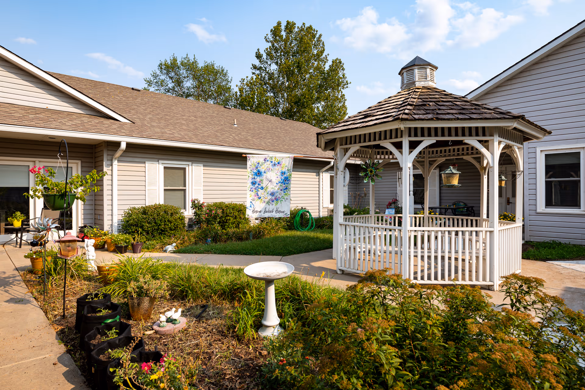 Outdoor garden area at Homestead Assisted Living of Derby featuring a white wooden gazebo with a shingled roof, surrounded by plants and flowers. The building exterior is beige with white trim, and there is a decorative flag with floral design and the words 'Home Sweet Home' hanging near the windows.