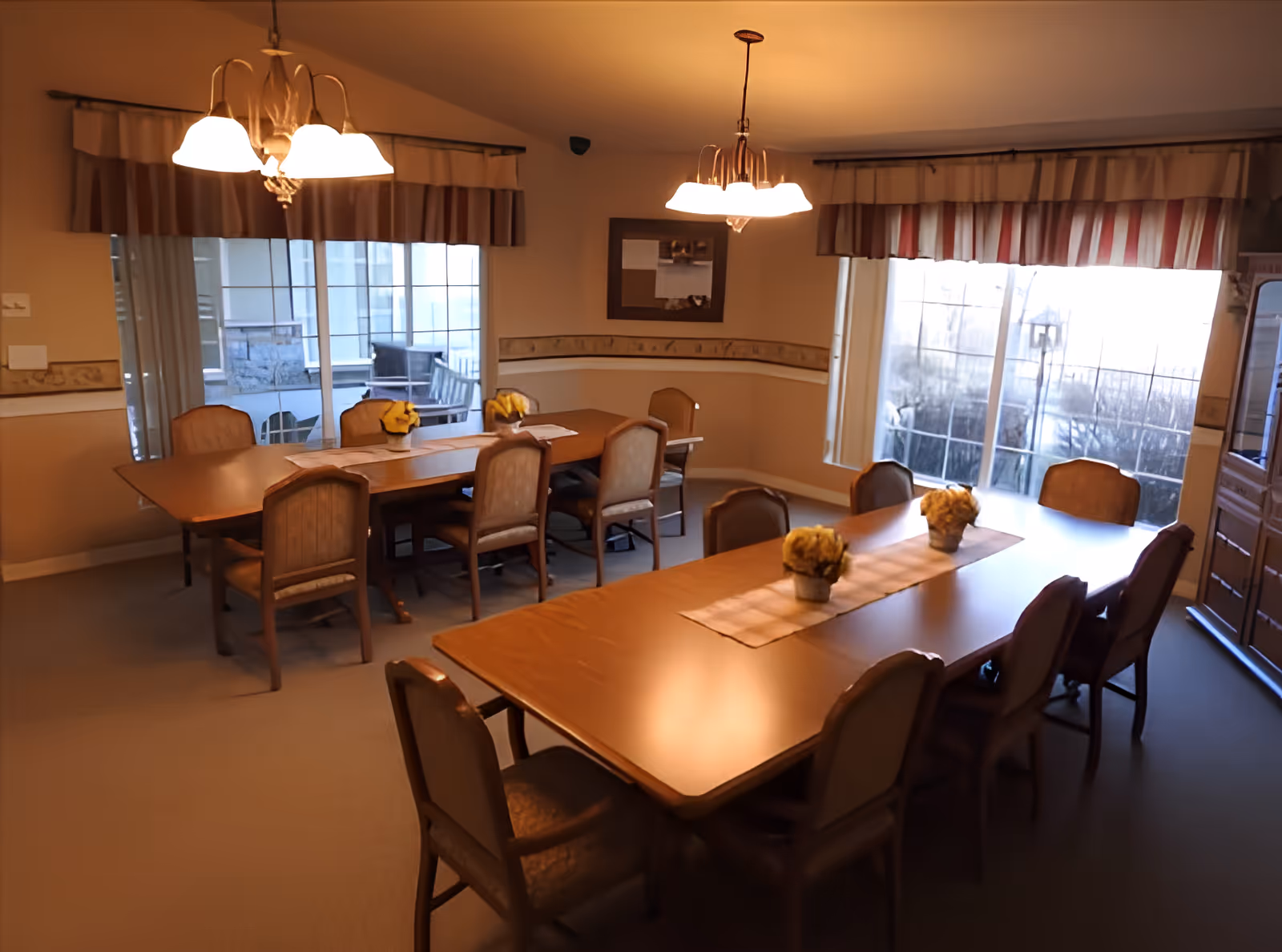 A communal dining room with two long wooden tables, upholstered chairs, hanging light fixtures, and large windows.