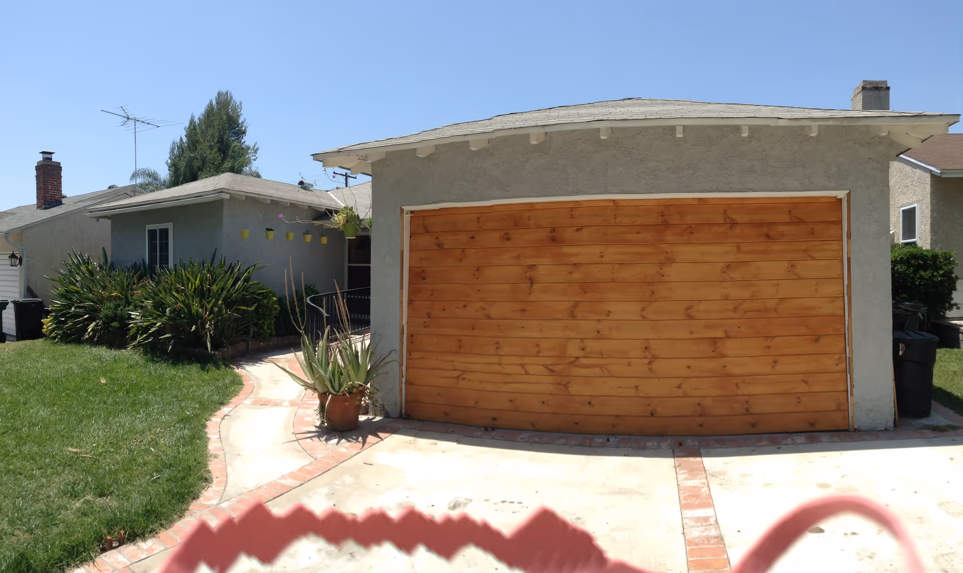 Front exterior of a single-story house showing a wooden garage door, driveway, lawn, and potted plants.