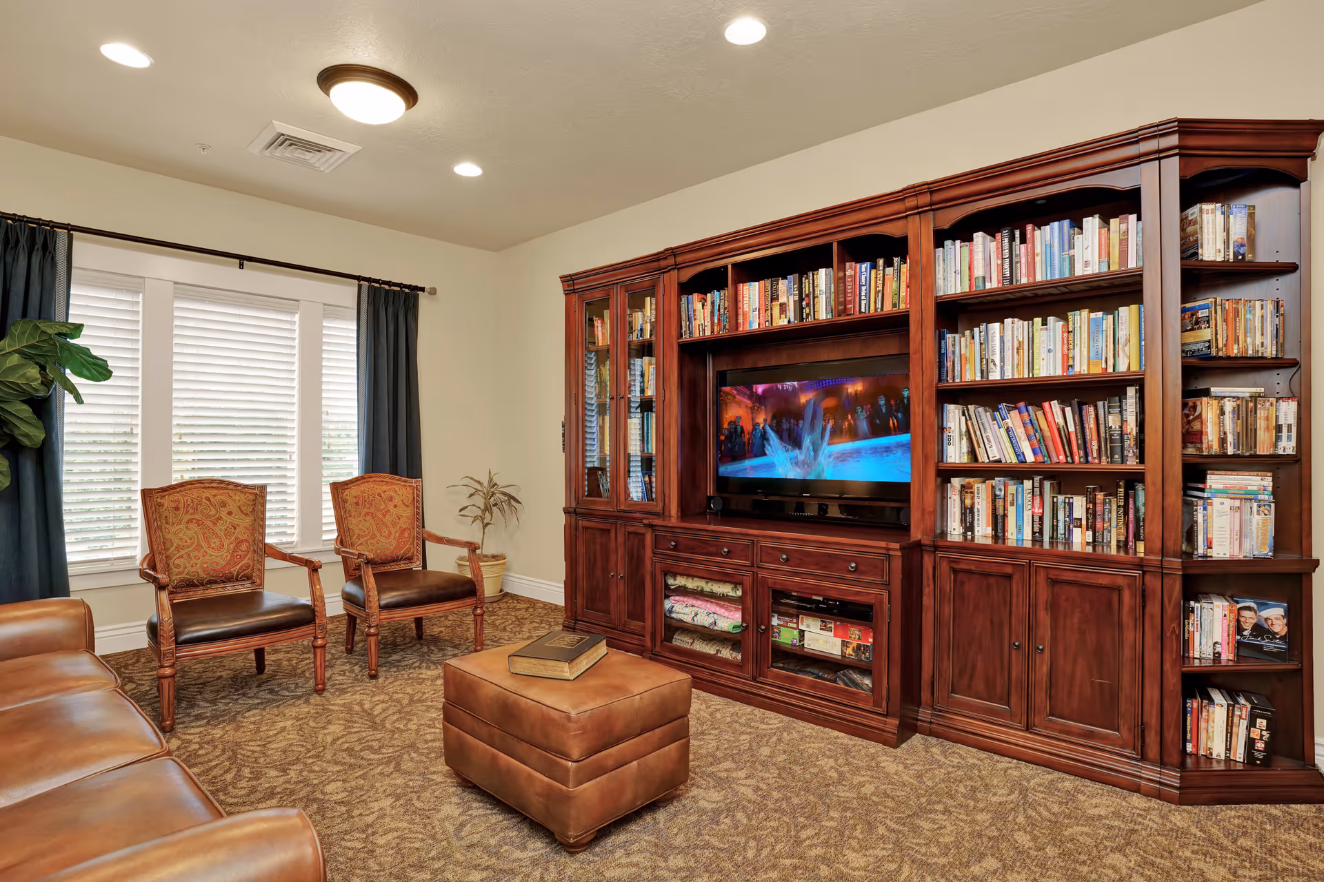 A cozy living room with a large wooden bookshelf filled with books and a flat-screen TV in the center. The room has a brown leather sofa, two upholstered armchairs with wooden frames, a brown leather ottoman with a book on top, and a window with white blinds and dark curtains. The carpet has a patterned design, and there is a potted plant near the window.