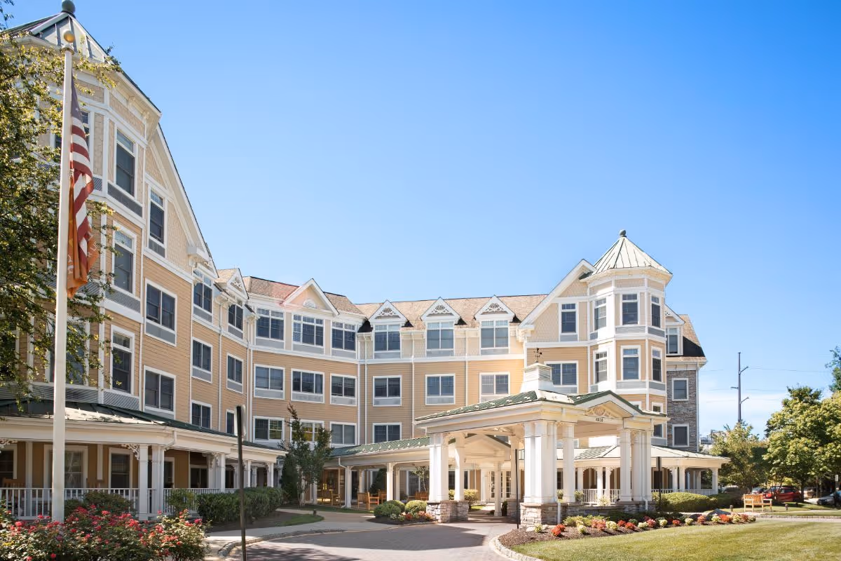 Exterior view of a large, multi-story senior living facility building with beige siding, white trim, and multiple windows. The building has a covered entrance with white columns and a green roof. There is a flagpole with an American flag in front, along with landscaped bushes and flowers under a clear blue sky.
