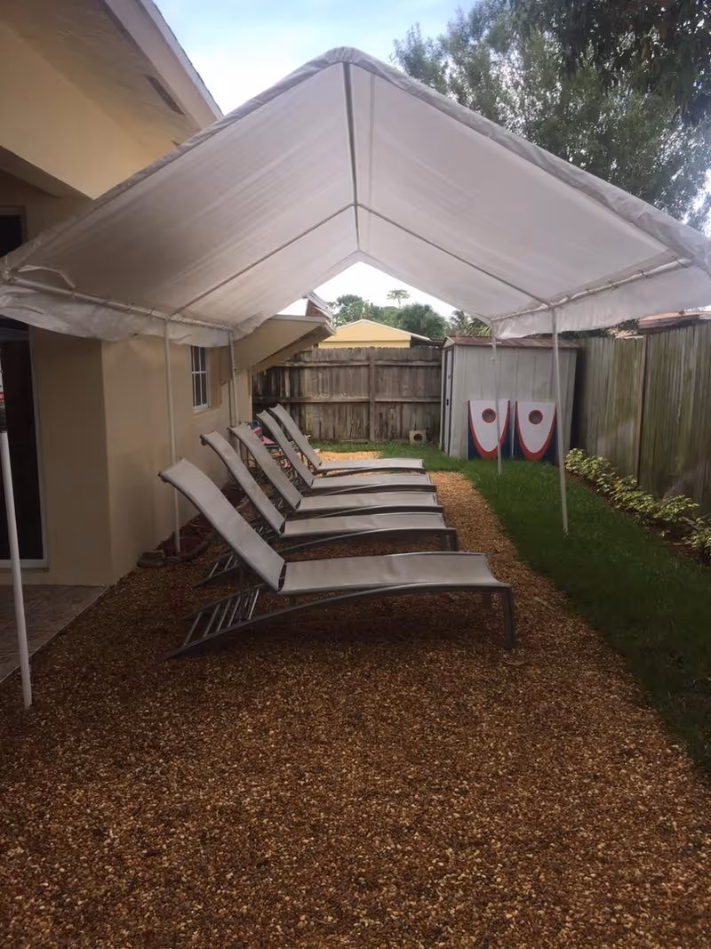 A row of reclining lounge chairs arranged under a white canopy in a narrow backyard alongside a house and wooden fence.