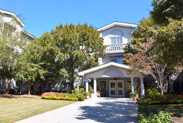 Front entrance of a multi-story senior living building with a covered portico, trees, and landscaped walkway.