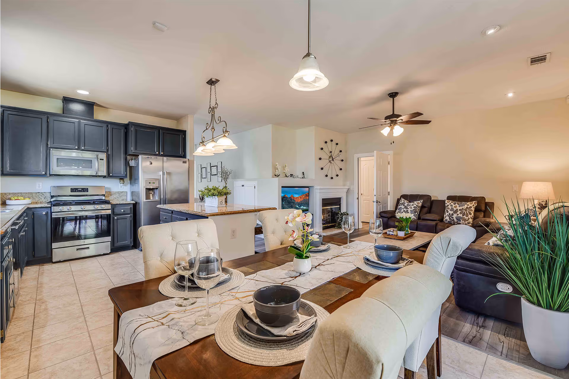 Open concept kitchen and living room area with a dining table set for four. The kitchen features dark cabinets, stainless steel appliances including a refrigerator and stove, and a central island with a plant on top. The living room has a dark leather sofa, a coffee table with plants, a ceiling fan with lights, and a fireplace with a TV mounted above it. The space is well-lit with pendant lights over the dining table and recessed lighting in the ceiling.