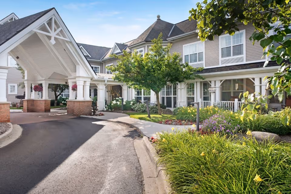 Exterior view of Sunrise of West Bloomfield senior living facility showing a covered entrance with white pillars and brick bases, surrounded by well-maintained landscaping including green trees, bushes, and colorful flowers under a clear blue sky.