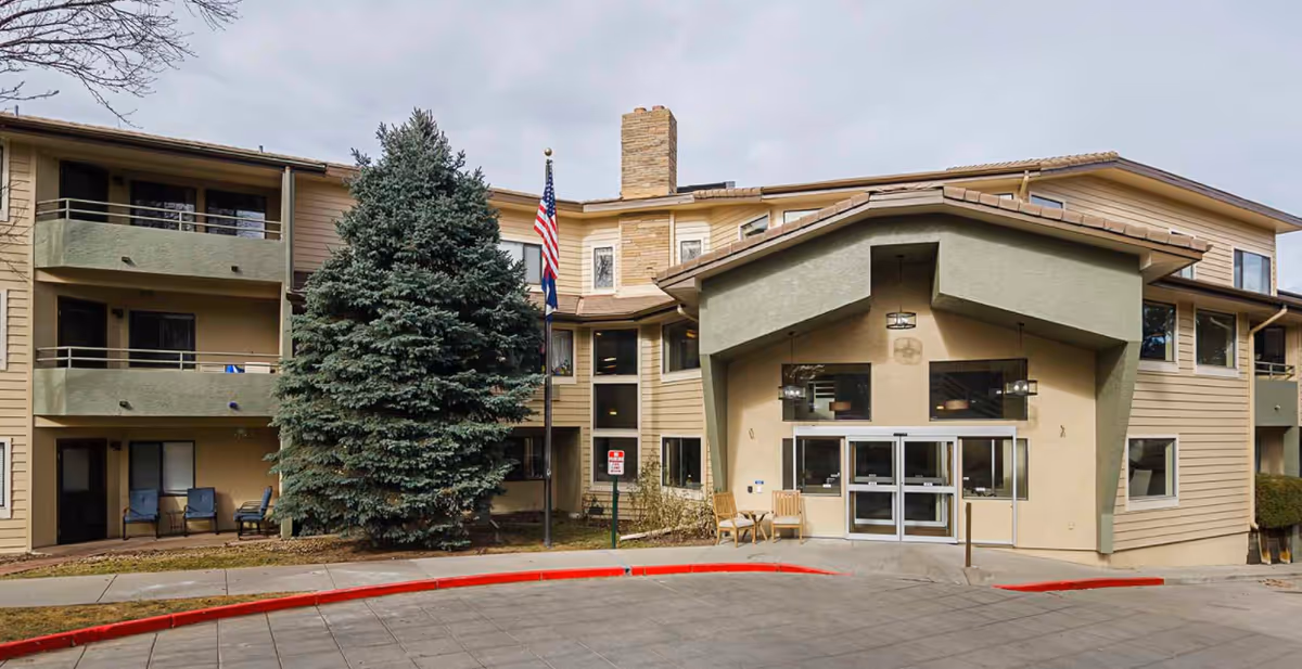 Exterior view of a multi-story senior living facility building with beige and green walls, a large evergreen tree near the entrance, two flagpoles with American and state flags, and a covered main entrance with glass doors. There are chairs placed near the entrance and a red curb along the driveway.