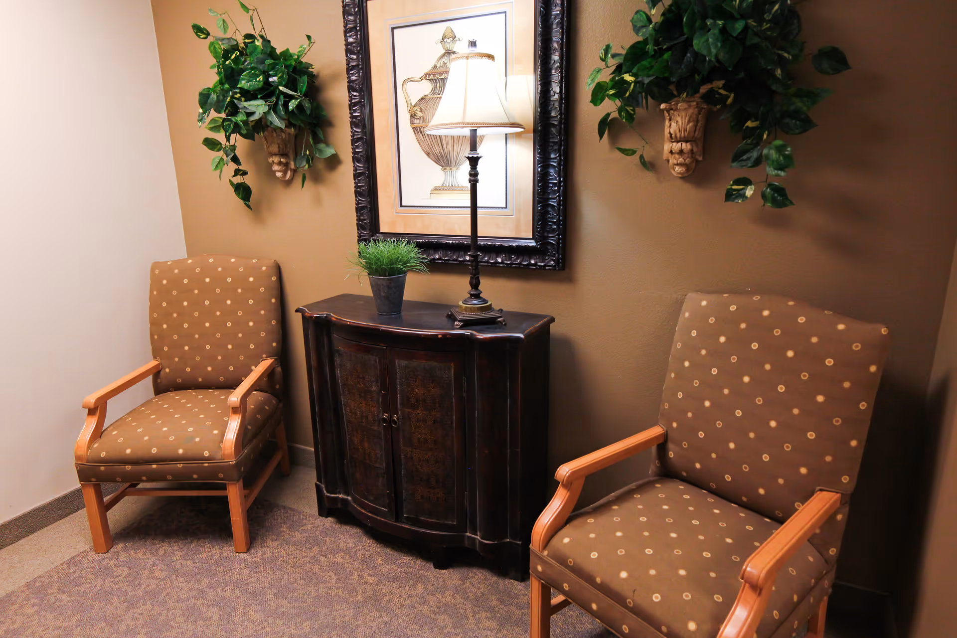 Two patterned armchairs flank a small dark wooden cabinet topped with a lamp, framed art, and wall plants in a cozy seating area.