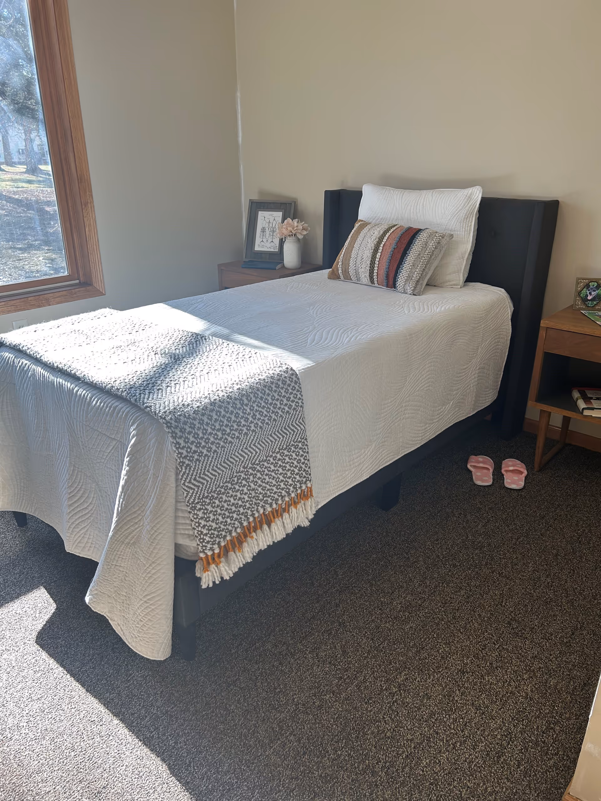 A small, neatly made single bed with a white quilt and a decorative throw blanket at the foot. There are two pillows, one plain white and one with a striped pattern. The bed is positioned next to a window with sunlight streaming in. On either side of the bed are wooden nightstands, one with a framed picture and a vase with flowers, and the other with a small framed photo and some books underneath. A pair of pink slippers is placed on the carpeted floor next to the bed.