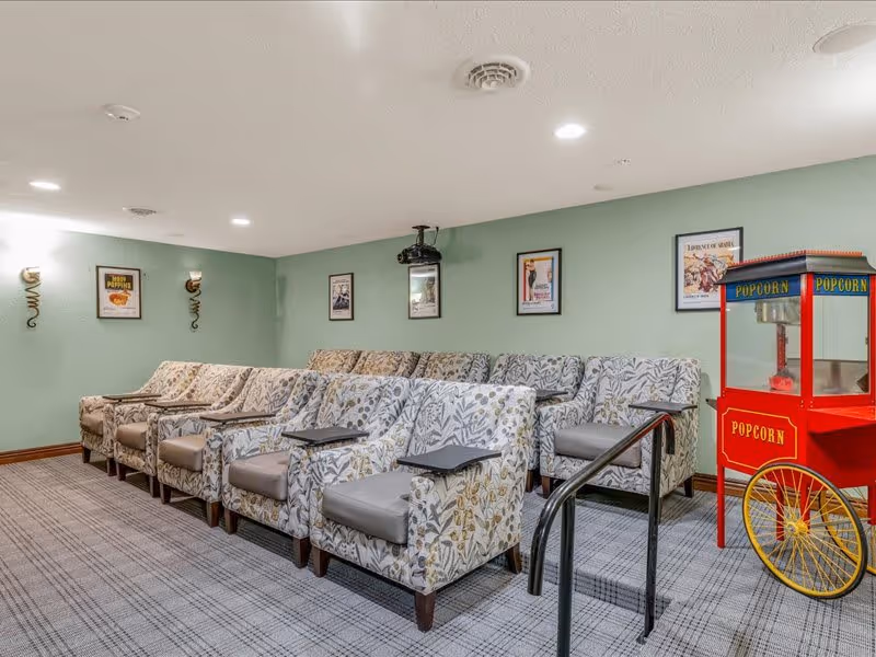 A small theater room with two rows of cushioned armchairs featuring floral patterns. Each chair has a small black tray attached. The walls are painted light green and decorated with framed movie posters. A red popcorn machine with yellow wheels is positioned on the right side of the room. The floor is carpeted with a checkered pattern.